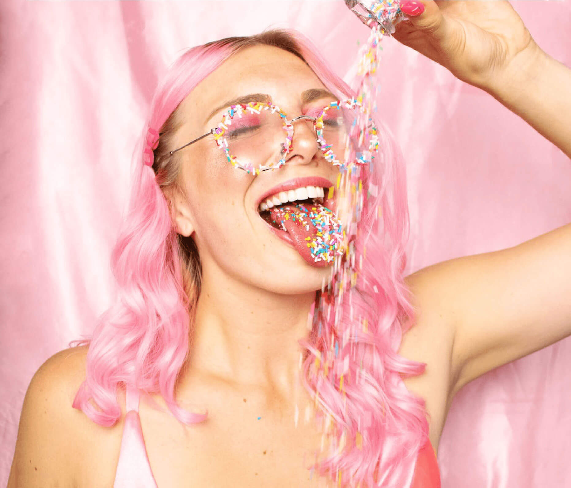 Young woman with pastel pink hair and glasses wearing rainbow sprinkles, embodying the playful spirit of BlissVixen's premium toys and unforgettable parties
